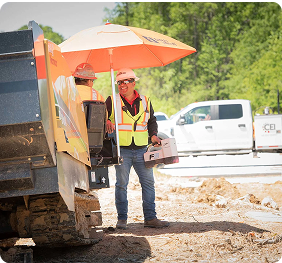 Construction workers in safety vests at a job site, one holding an orange umbrella next to heavy equipment.