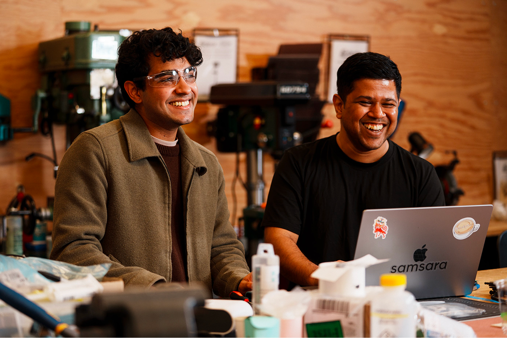 Two smiling people in a workshop, one wearing safety glasses, looking at a laptop with "samsara" sticker.