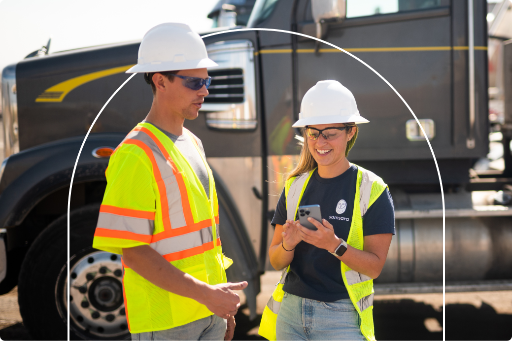 Two construction workers in hard hats and safety vests reviewing information on a smartphone near a truck.