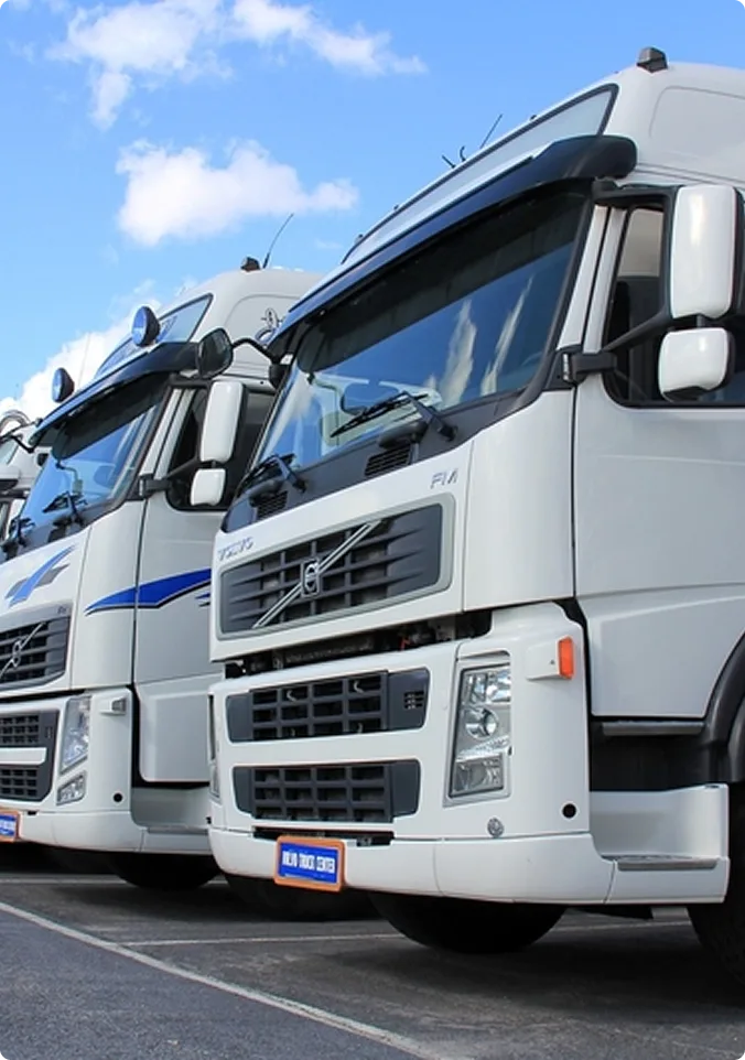 Three maroon semi trucks parked in a row at loading docks beneath a blue warehouse building at dusk.