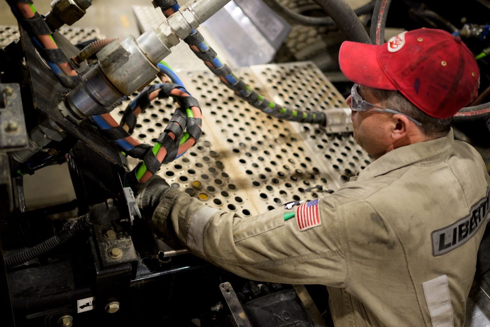 Worker in red cap and Liberty uniform with American flag patch operating industrial machinery with colorful cables.