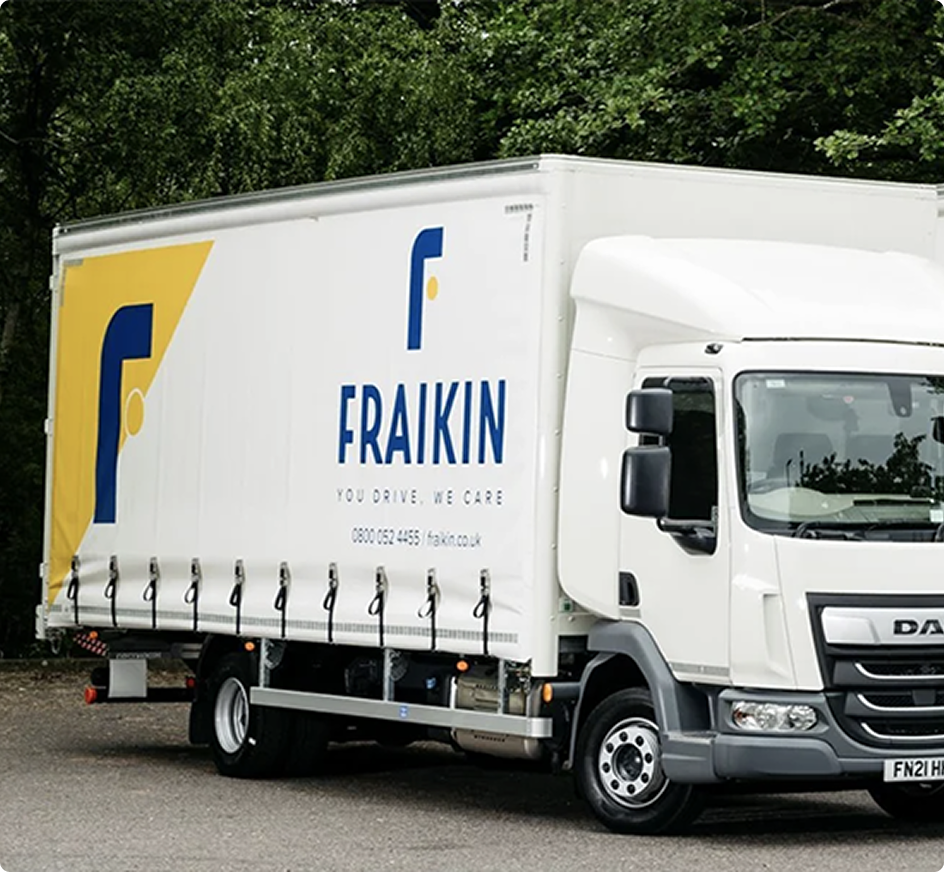 White Fraikin delivery truck with blue and yellow logo parked on a road with green trees in the background.