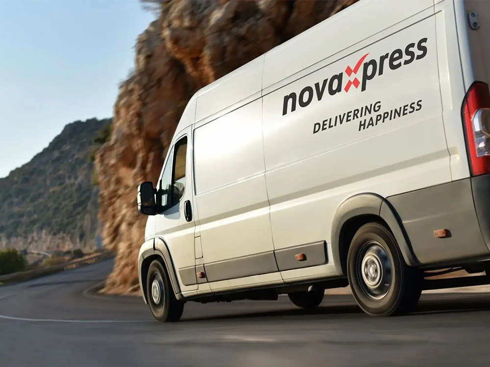 A white NovaXpress delivery van with "Delivering Happiness" text driving on a road beside a rocky cliff.