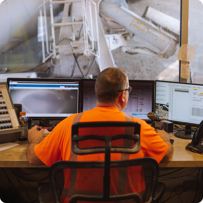 Persoon in oranje shirt bewaakt industriële apparatuur via meerdere computerschermen in een controlekamer.