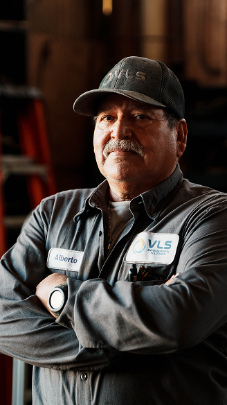 Alberto, a VLS worker, stands with arms crossed wearing a gray uniform and cap, looking confidently at the camera.