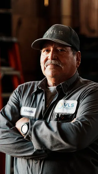 Alberto, a VLS worker, stands with arms crossed wearing a gray uniform and cap, looking confidently at the camera.