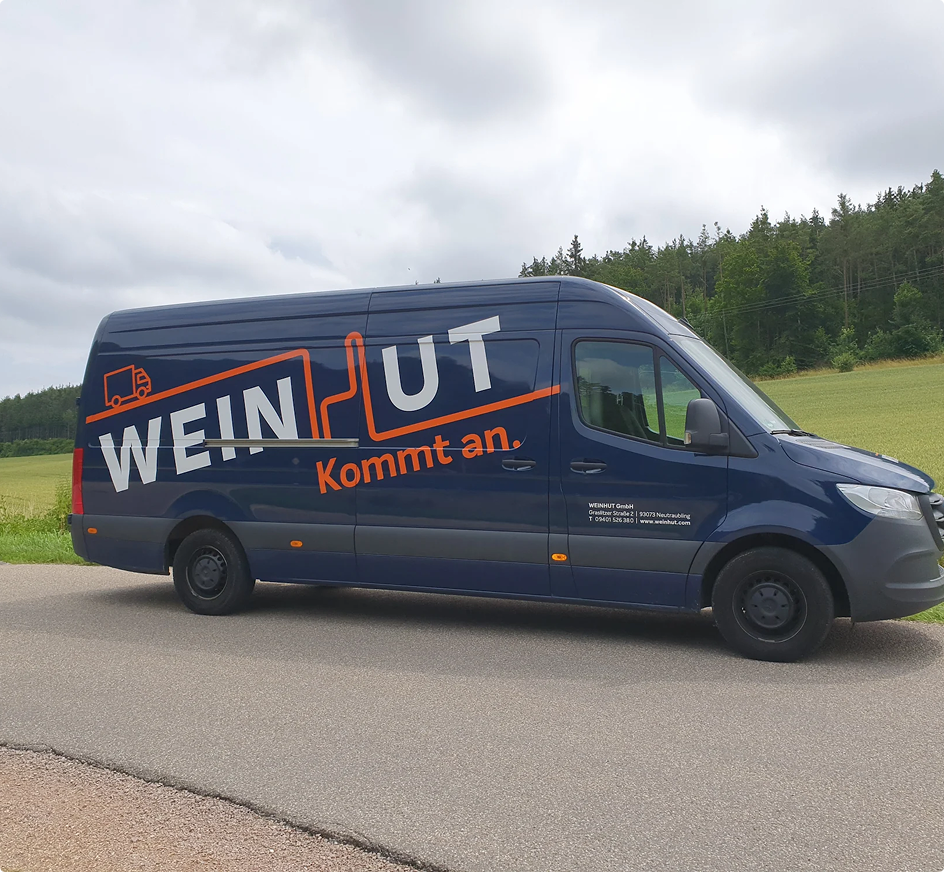 Blauer Lieferwagen mit "WEIN UT kommt an" Aufschrift auf einer Landstraße vor grünem Wald und Feldern unter bewölktem Himmel.