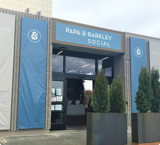 Storefront of Papa & Barkley Social with blue signage and banners, flanked by small evergreen trees at the entrance.