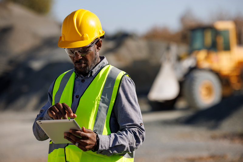 A construction worker in a yellow hard hat and hi-vis vest uses a tablet on a building site with heavy machinery in the background.