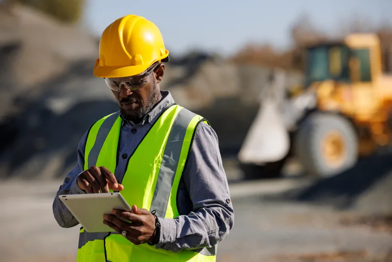 A construction worker in a yellow hard hat and hi-vis vest uses a tablet on a building site with heavy machinery in the background.