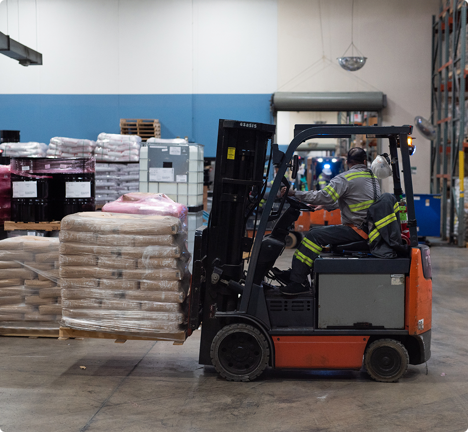 Worker operating a forklift carrying a pallet of wrapped goods in a warehouse with stacked inventory.