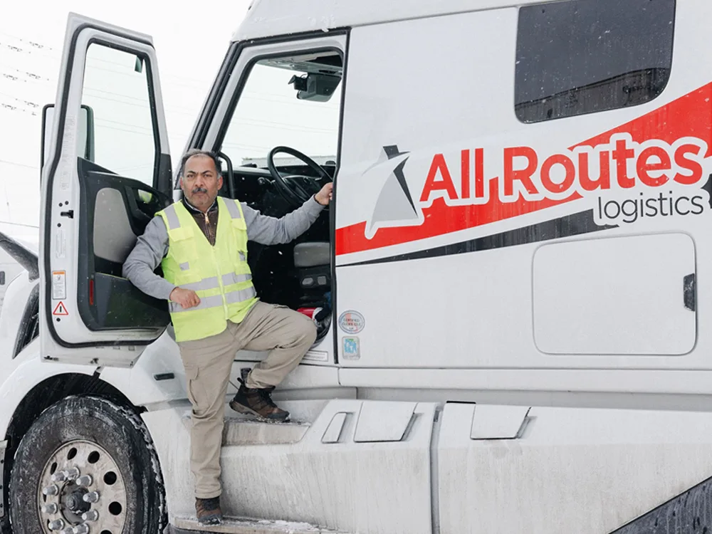 Driver in reflective yellow vest standing outside the door of an All Routes Logistics truck in the winter.