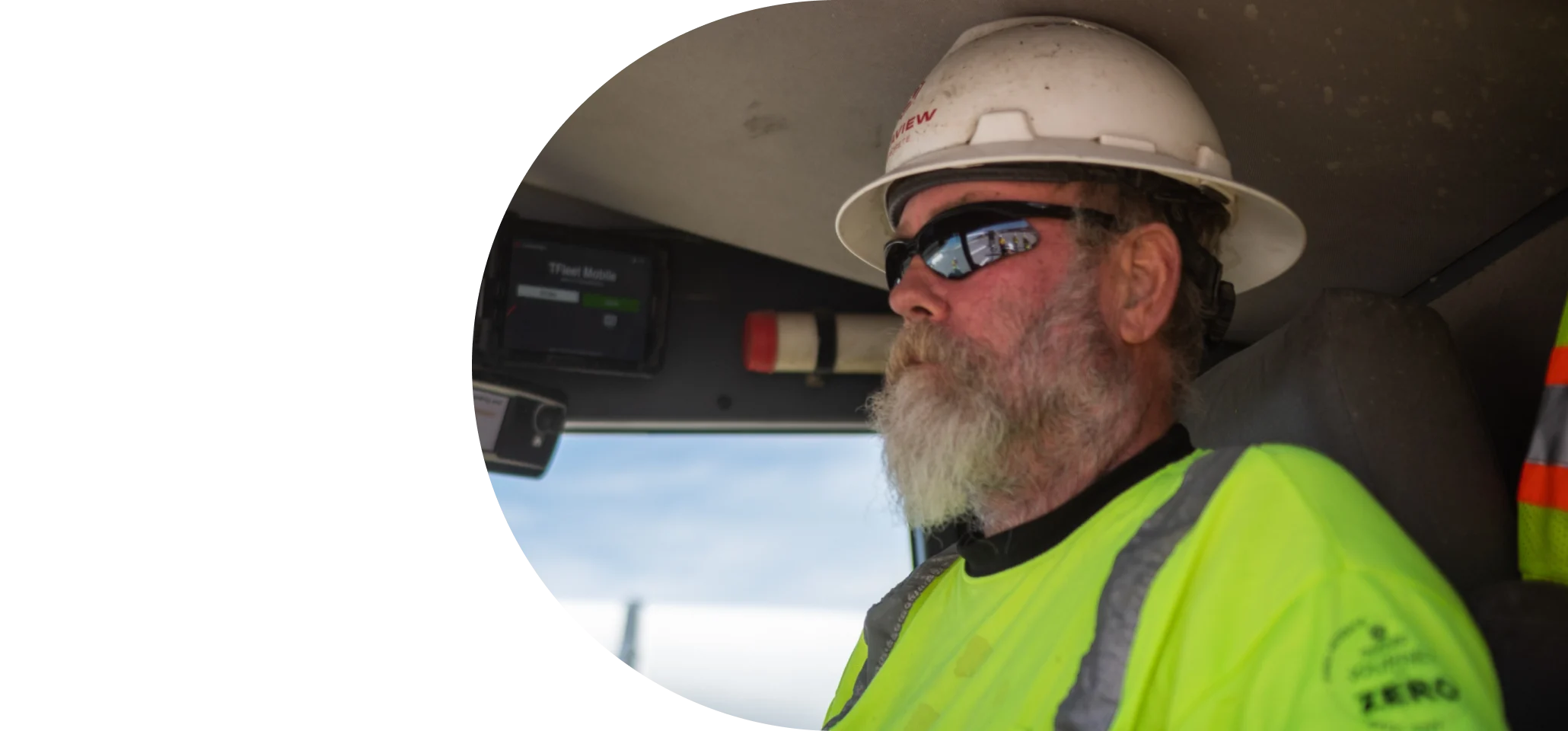 A man with a beard, wearing a hard hat, sunglasses, and a high-visibility shirt, driving a truck