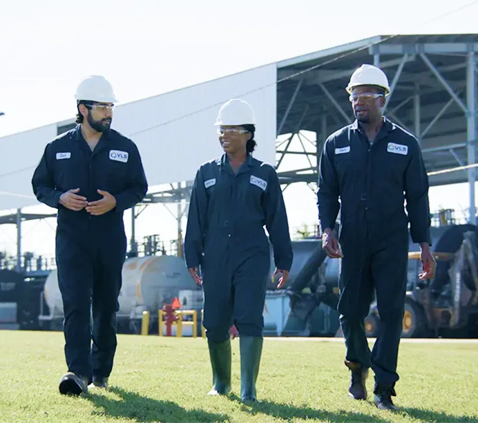 Three industrial workers in navy coveralls and white hard hats walking across grass at a construction or plant site.
