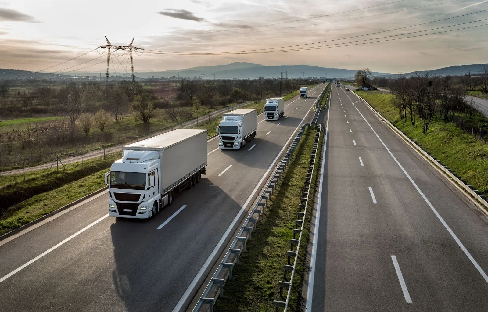 White lorries travelling on a motorway with mountains in the distance under an overcast sky, power lines visible.