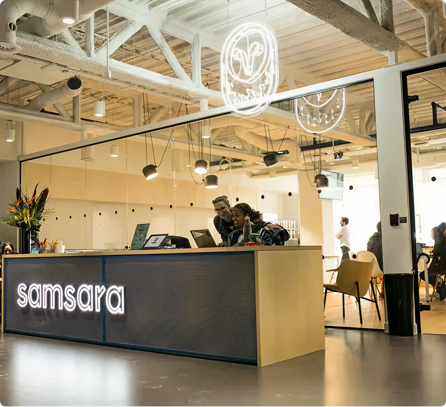 Modern interior with "samsara" illuminated reception desk, pendant lights, and neon logo sign on industrial ceiling.