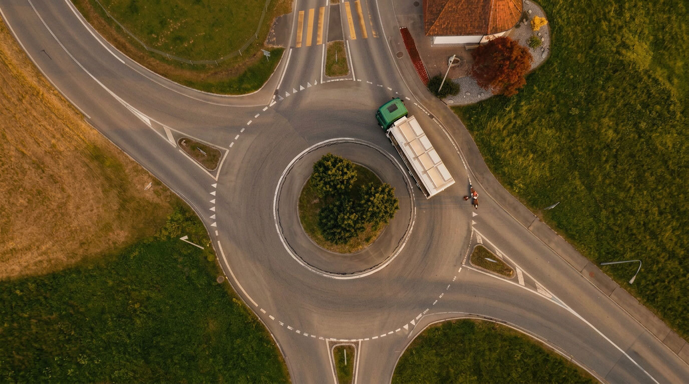 Luftaufnahme eines Kreisverkehrs mit einem grünen LKW, umgeben von grünen Wiesen und einem Gebäude.
