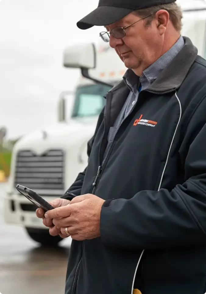 Older man in black jacket and cap checking smartphone with semi-truck visible in background.