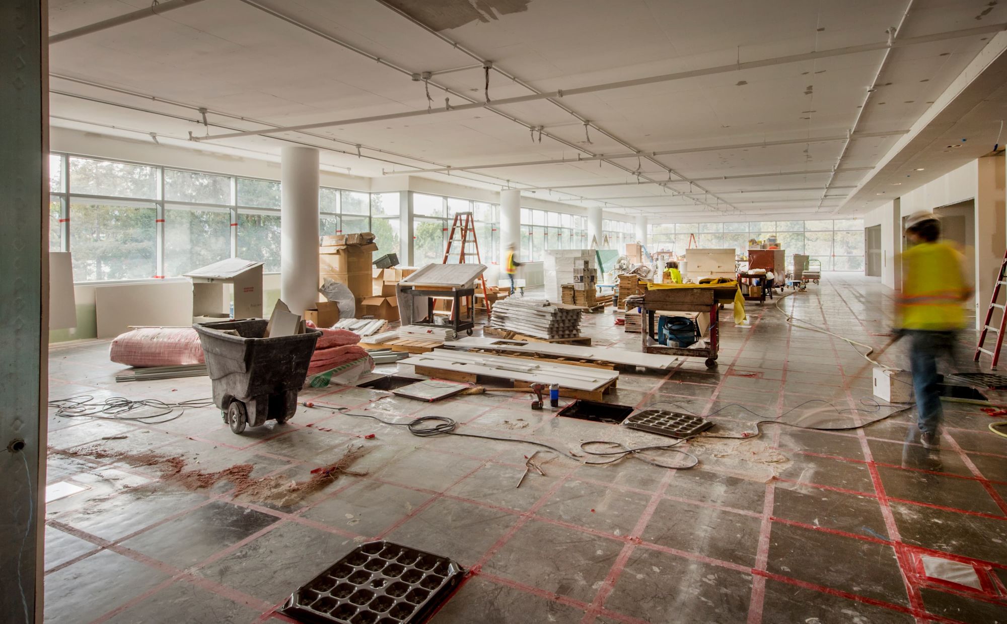 Großer Büroraum im Umbau mit Baumaterialien, Werkzeugen und Bauarbeiter in gelber Weste. Große Fensterfront mit Tageslicht.
