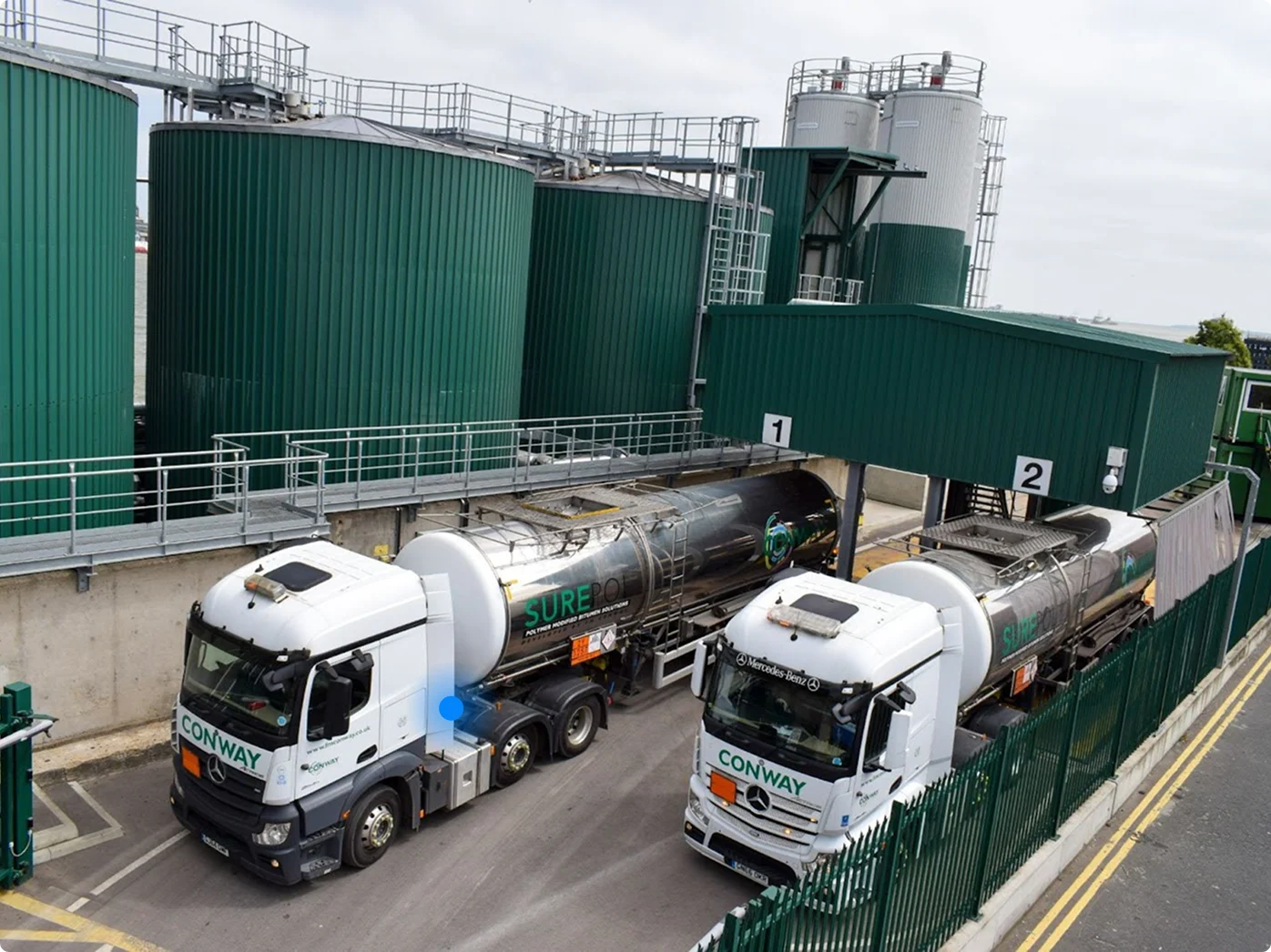 Two white Conway tanker trucks parked at a loading facility with large green storage tanks and processing equipment.