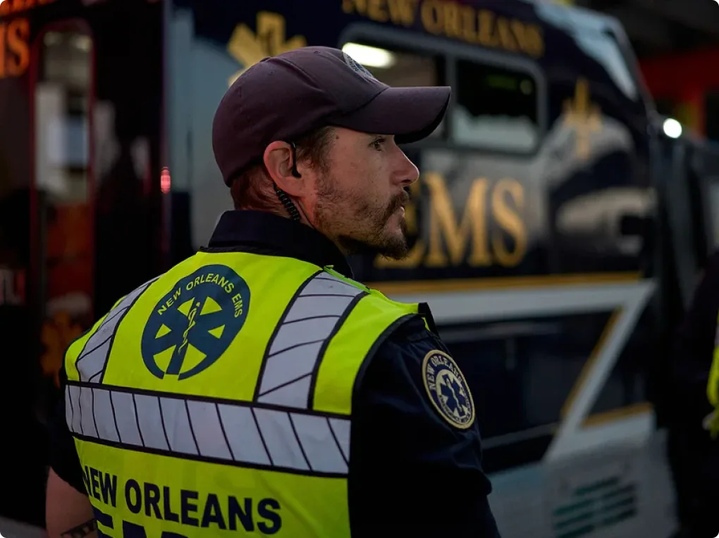 New Orleans EMS worker in high-visibility vest monitoring a Mardi Gras parade with colorful floats in the background.