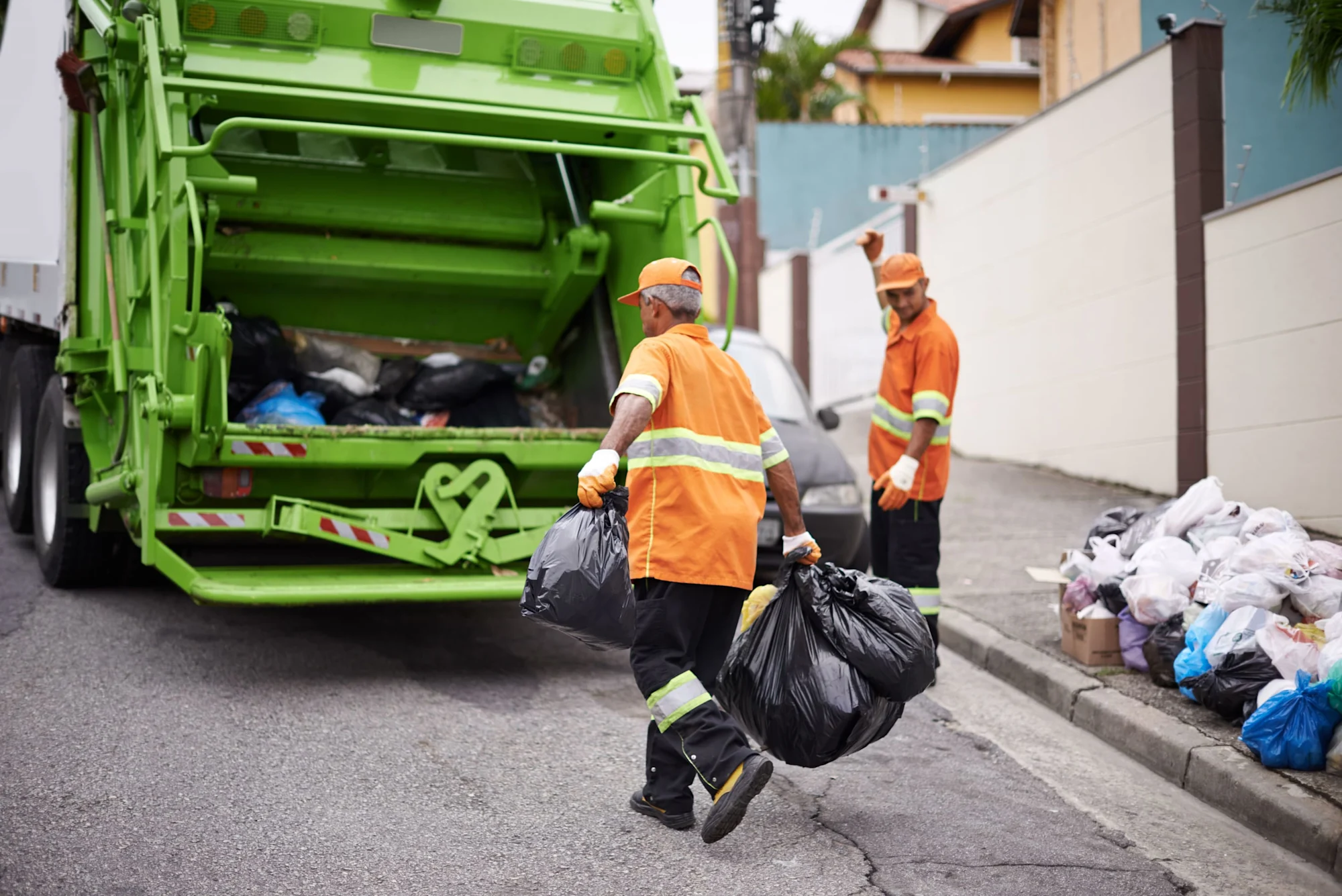Müllarbeiter in orangefarbener Kleidung laden schwarze Müllsäcke in einen grünen Müllwagen in einer Wohnstraße.