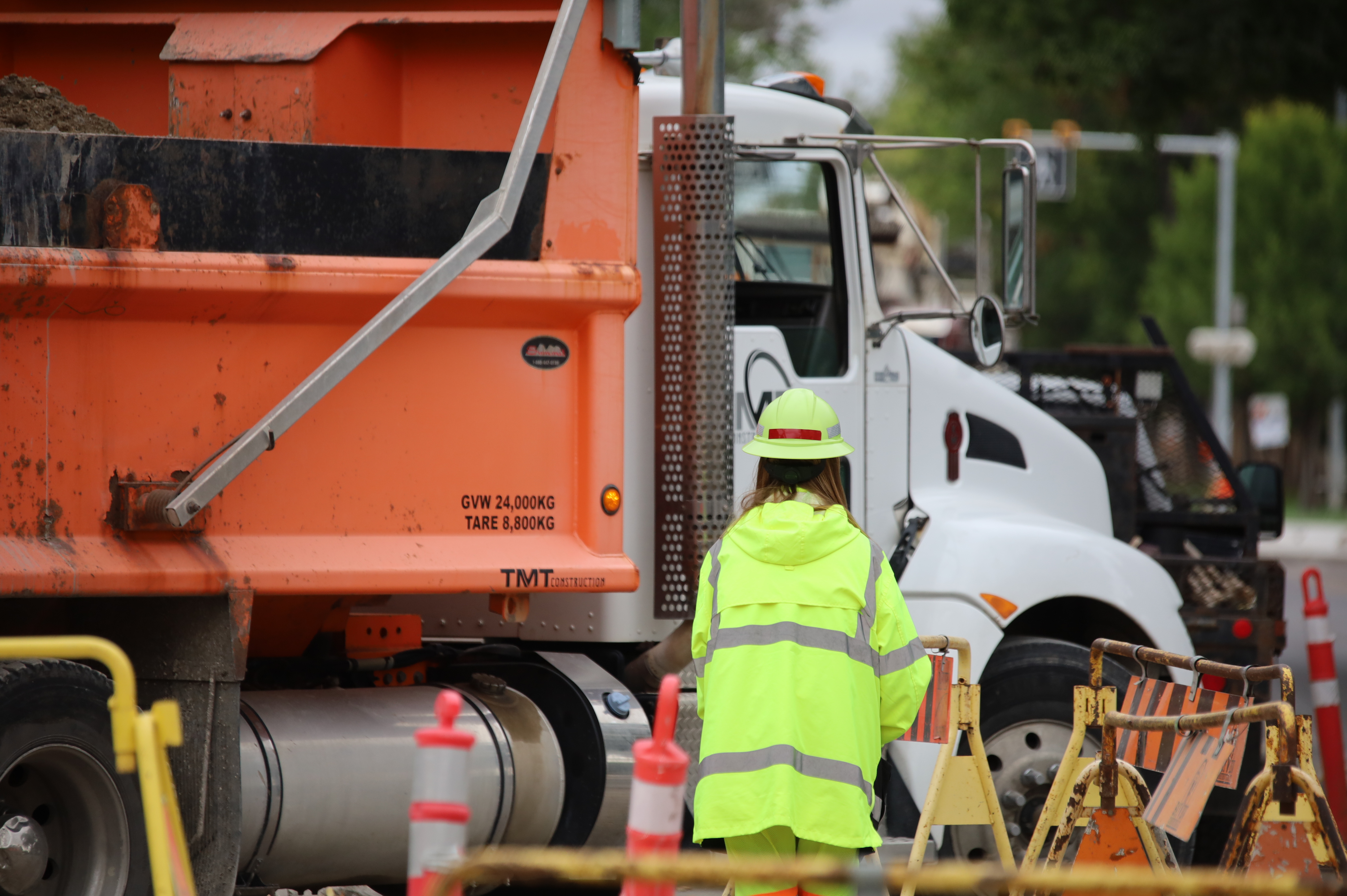 Construction worker in bright yellow safety gear standing near an orange dump truck at a roadwork site.