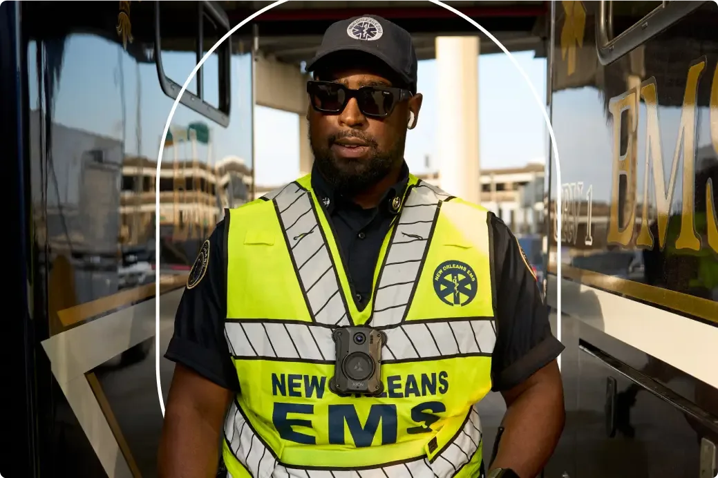 Emergency responder wearing neon yellow New Orleans EMS vest, black cap and sunglasses, standing near vehicles.