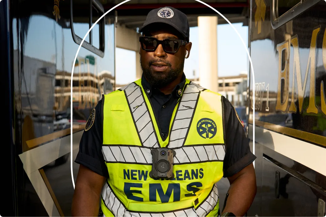 Un socorrista con un chaleco amarillo neón de New Orleans EMS, una gorra negra y gafas de sol, de pie cerca de los vehículos.