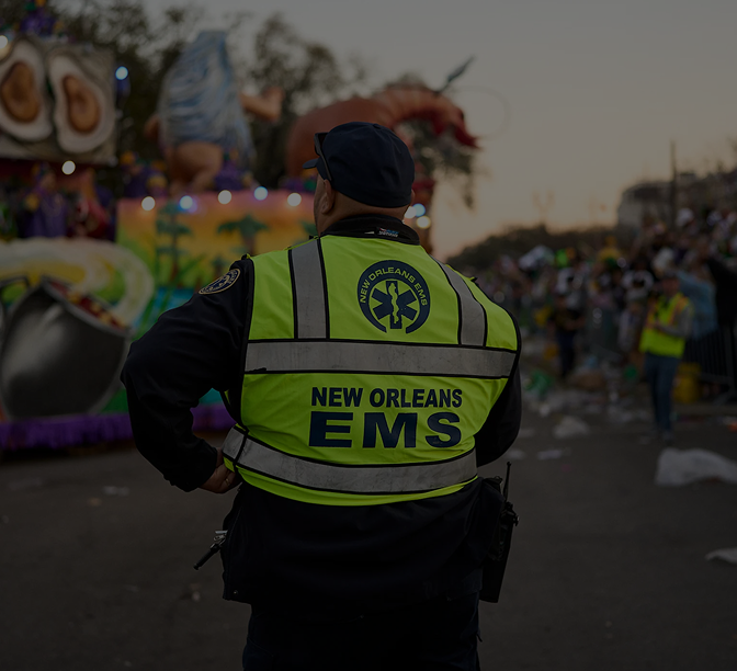 New Orleans EMS worker in bright yellow vest watching over a parade with colorful floats in the background.