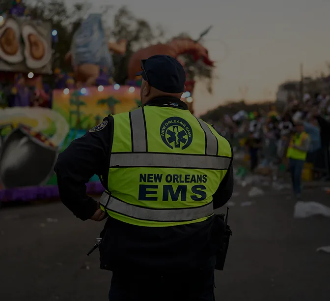 New Orleans EMS worker in bright yellow vest watching over a parade with colorful floats in the background.