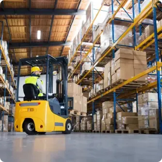 Worker operating a yellow forklift in a large warehouse with tall storage racks filled with stacked boxes and pallets.