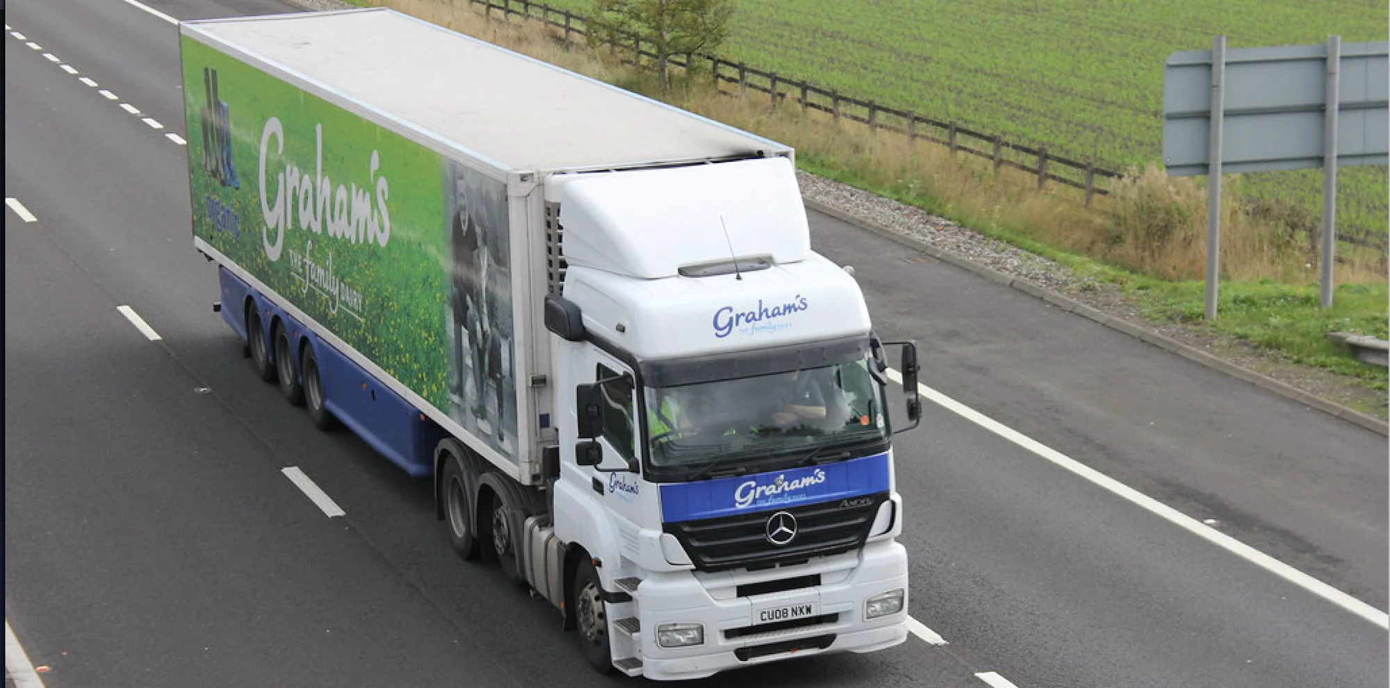 Camion de livraison Graham's blanc et bleu avec remorque verte circulant sur l'autoroute à côté d'un champ vert.