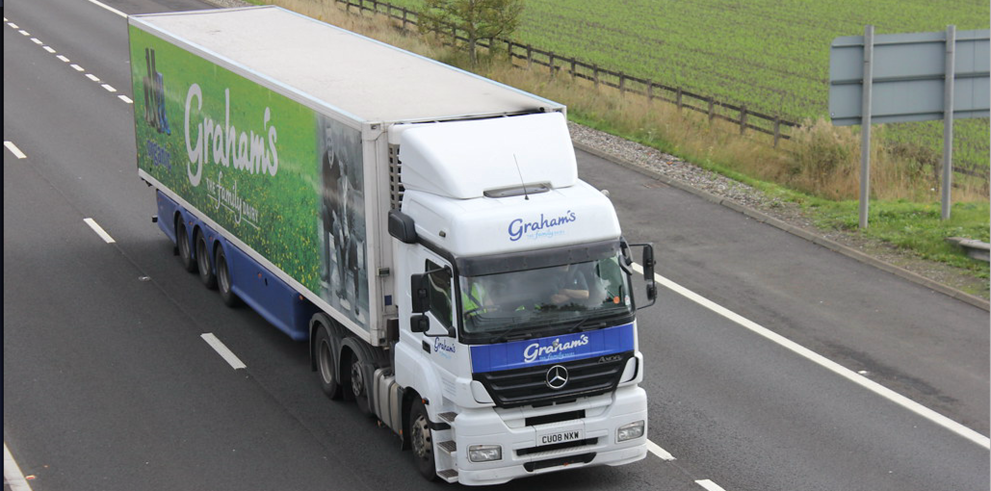 Witte Mercedes vrachtwagen van Graham's met groene en blauwe oplegger rijdt op de snelweg langs groen landschap.