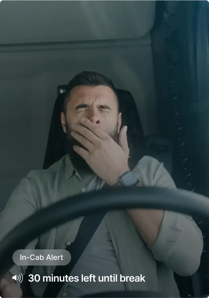 Tired driver yawning in truck cab with "In-Cab Alert" notification showing "30 minutes left until break"