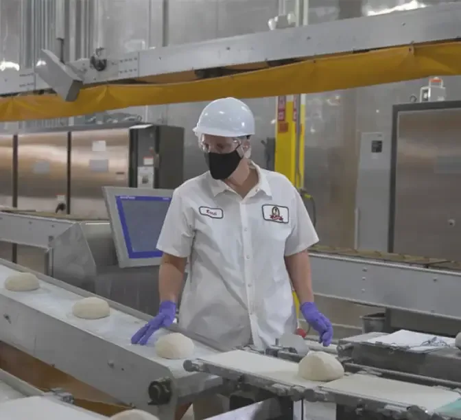 Worker in white hard hat and mask monitoring dough on a production line conveyor belt in a food factory.