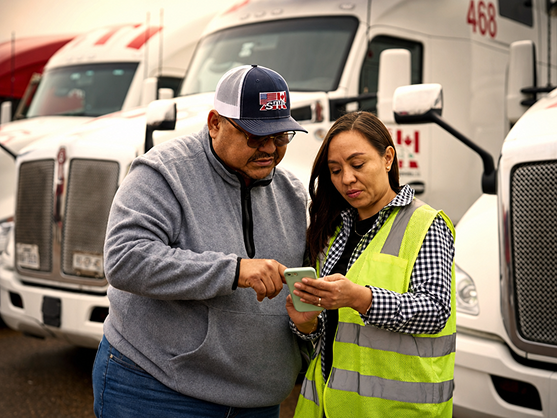 Two workers review a smartphone in a truck depot, with white semi-trucks parked behind them.