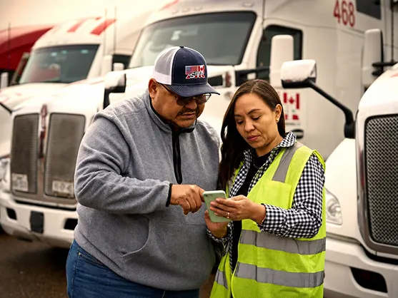 Two workers review a smartphone in a truck depot, with white semi-trucks parked behind them.