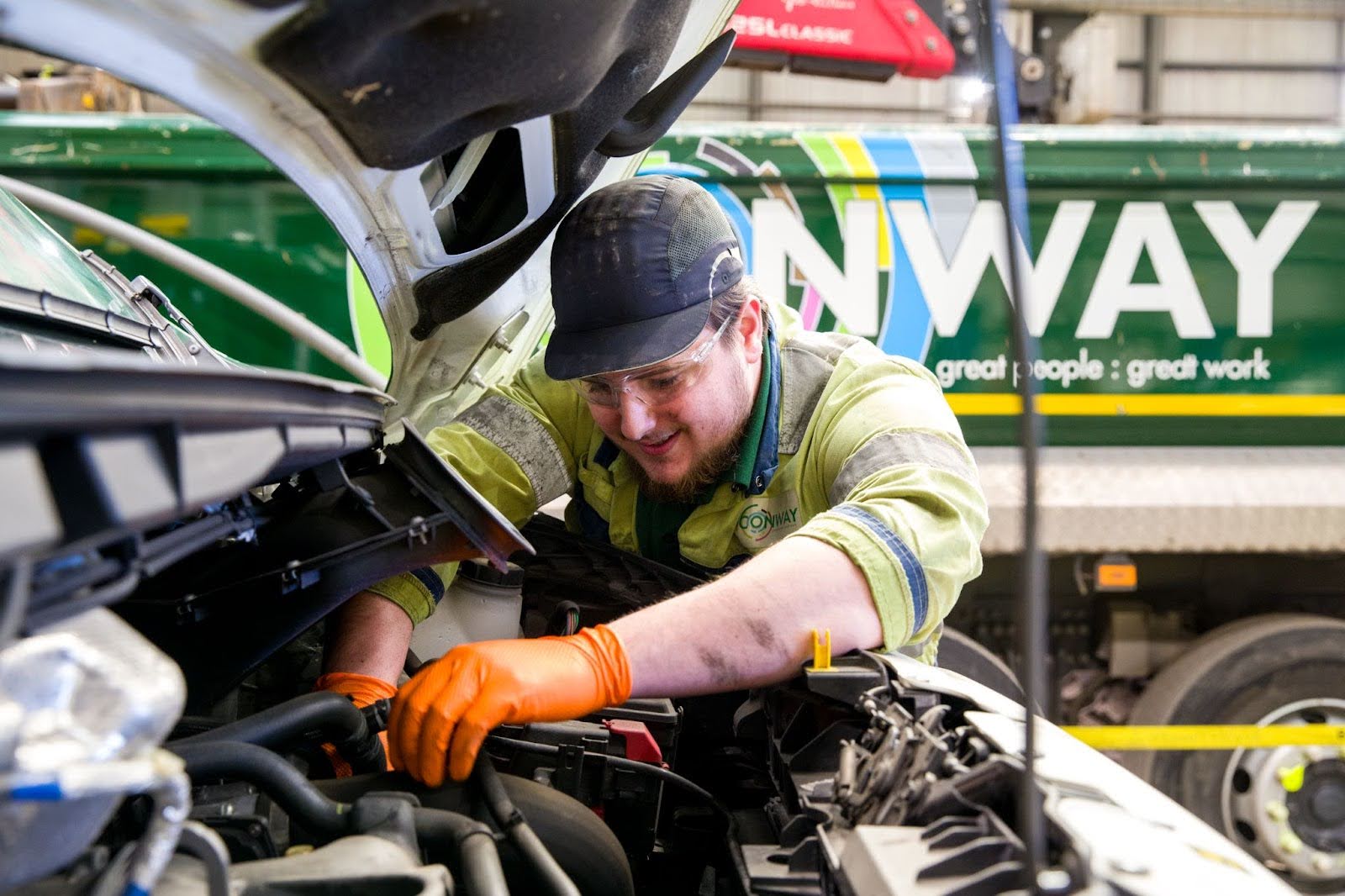 Mechanic in yellow uniform with orange gloves working on vehicle engine at Conway facility with green truck in background.