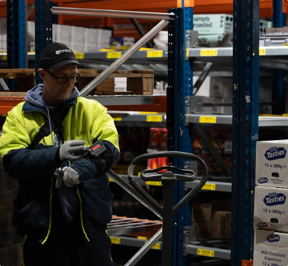 Travailleur en veste haute visibilité utilisant un scanner dans un entrepôt avec des étagères de stockage et des cartons.
