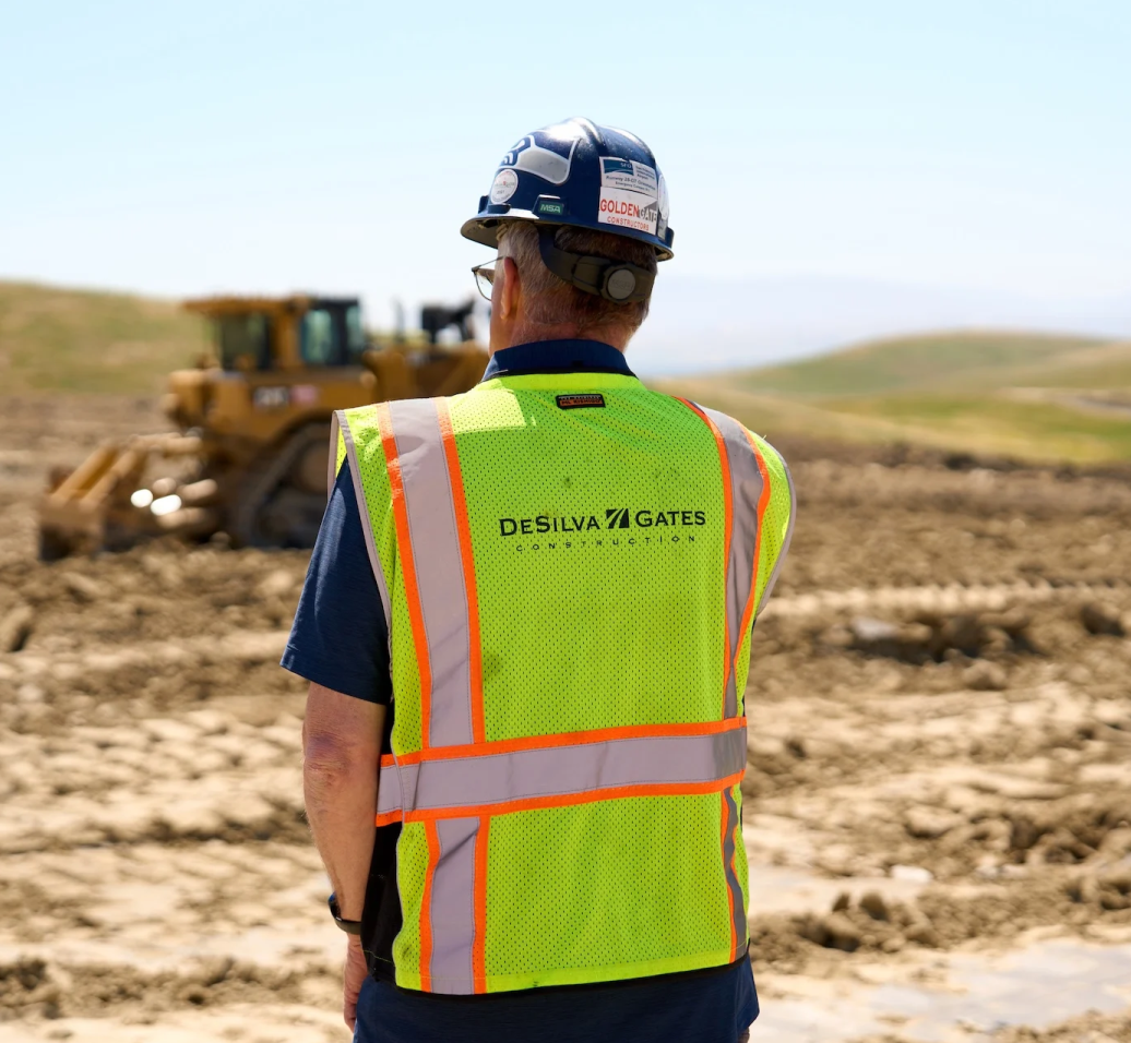 Construction worker in high-vis vest and hard hat overlooking bulldozer at earthworks site