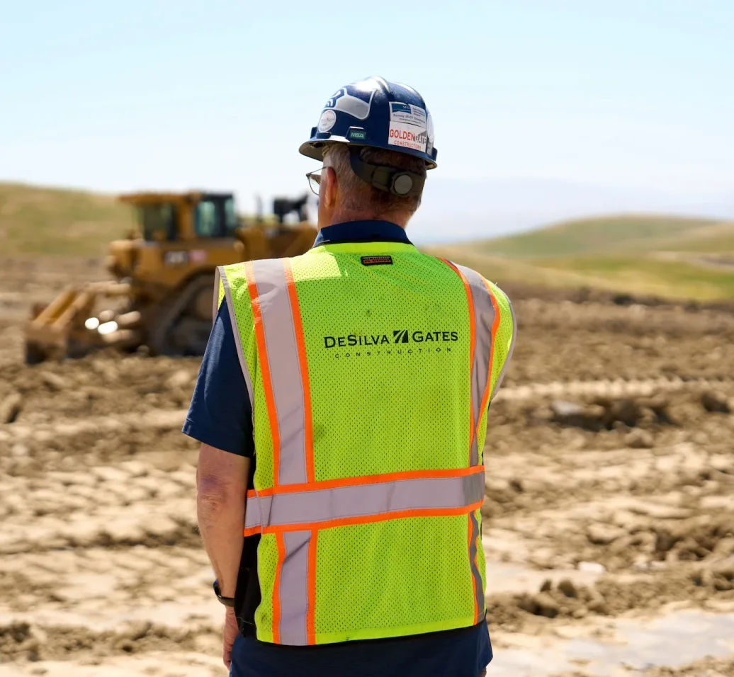 Construction worker in safety vest and hard hat overlooking bulldozer at earthwork site.
