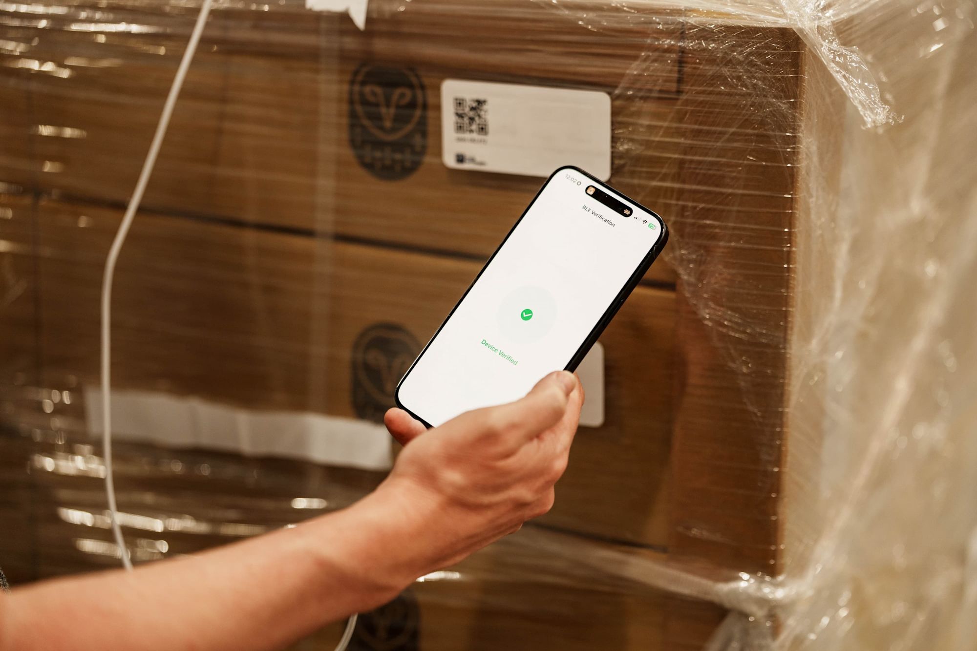 A hand holds a smartphone displaying "Device Verified" in green near a wrapped cardboard box with a QR code label.