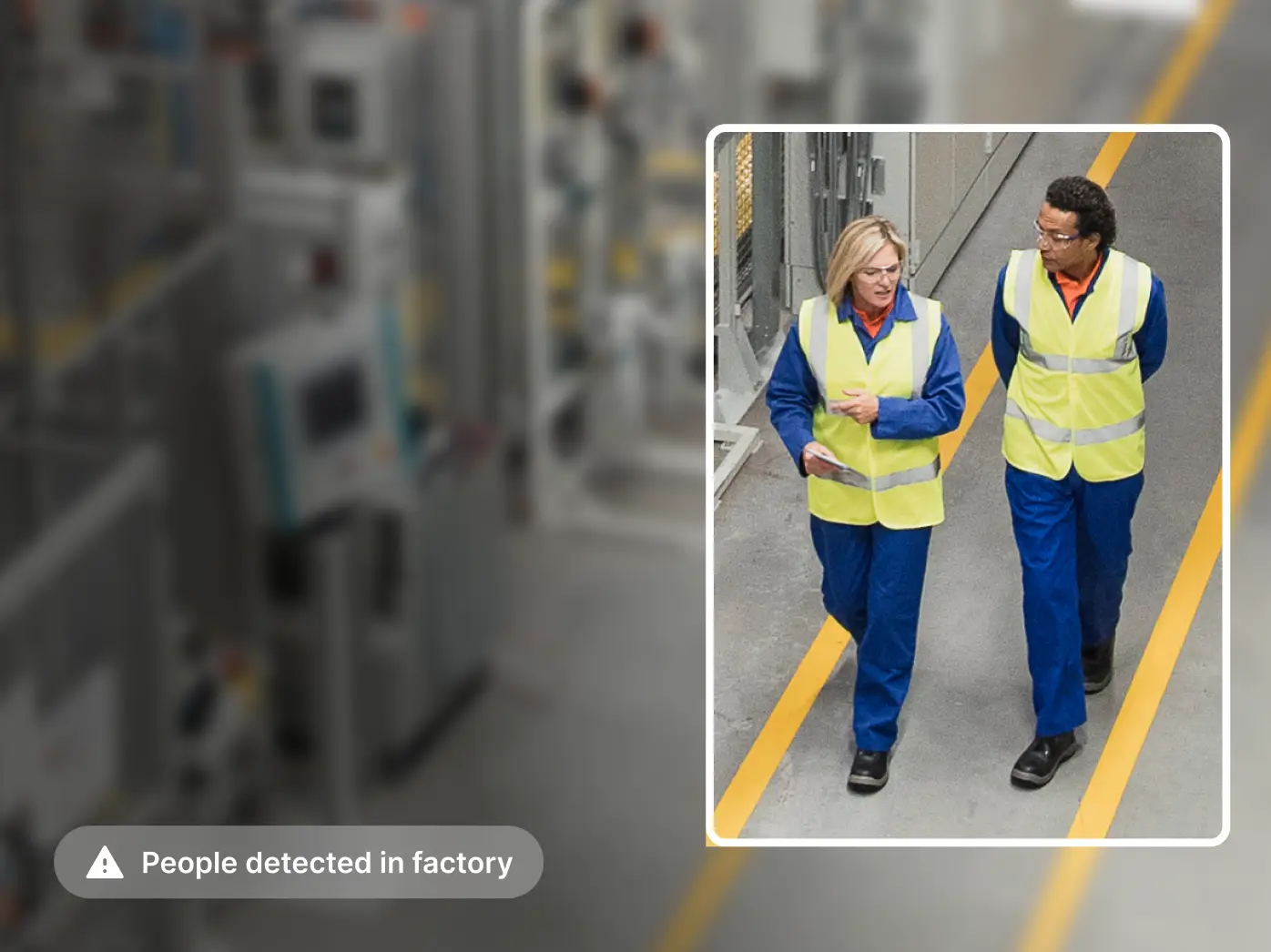 Two workers in blue uniforms and yellow safety vests walking along yellow floor markings in an industrial facility.