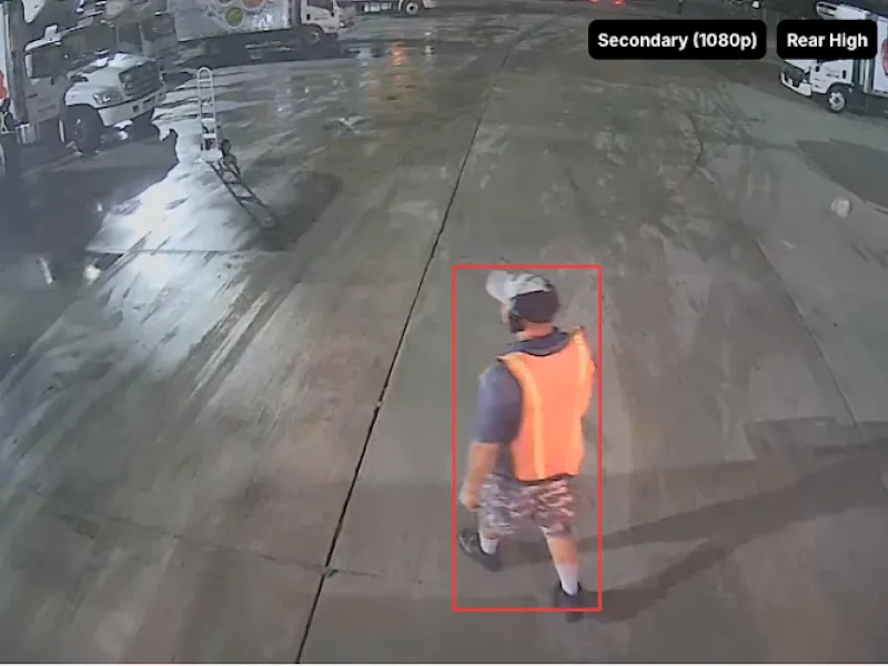 Persoon in oranje veiligheidsvest en camouflagebroek loopt over een nat parkeerterrein, vastgelegd door beveiligingscamera.