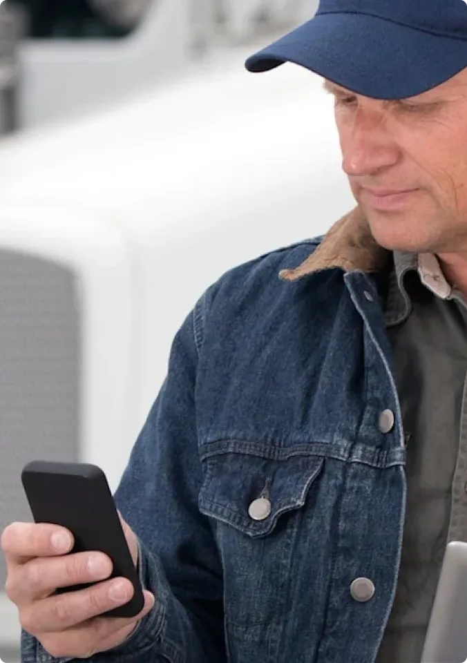 Person in blue cap and denim jacket looking at smartphone, with light background.