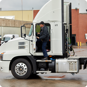 A person in dark clothing climbing into the cab of a white semi truck in a wet parking lot.