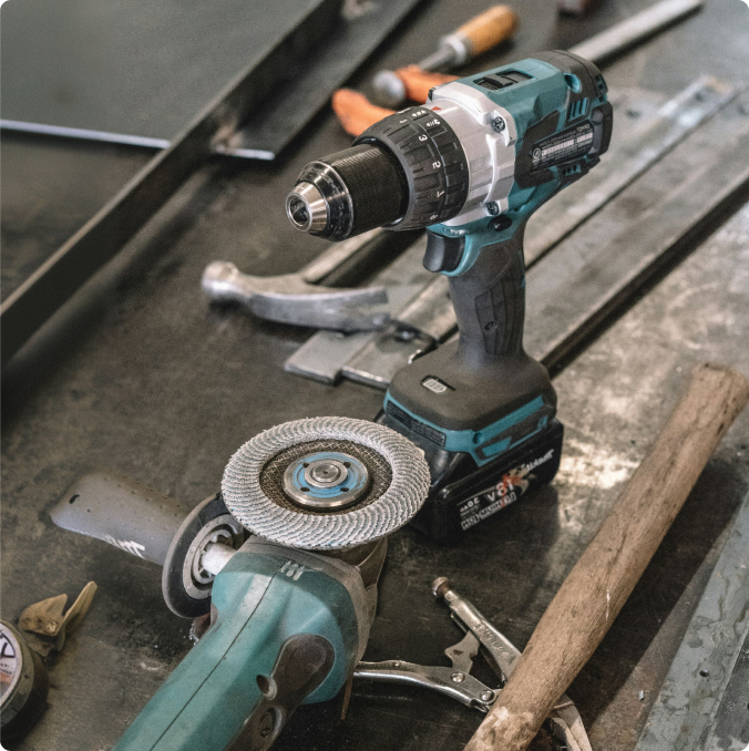 Teal power drill and angle grinder with metal grinding wheel on a workshop surface with various tools nearby.