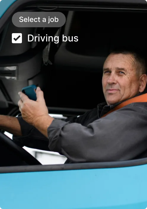 Bus driver in cab of blue bus with "Select a job" interface showing "Driving bus" option checked.