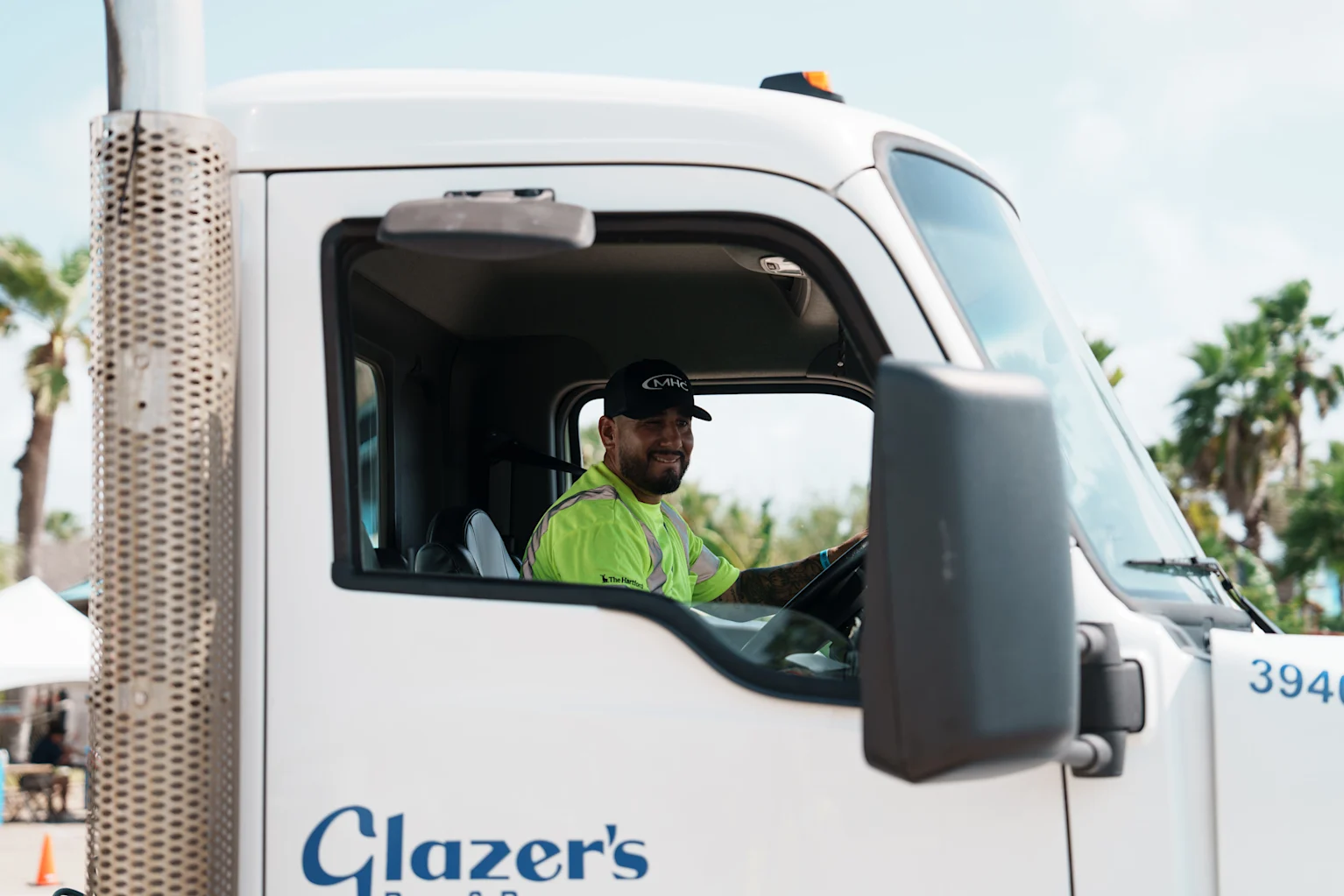 Chauffeur en gilet jaune fluorescent conduisant un camion blanc de l'entreprise Glazer's sous un ciel ensoleillé avec des palmiers.